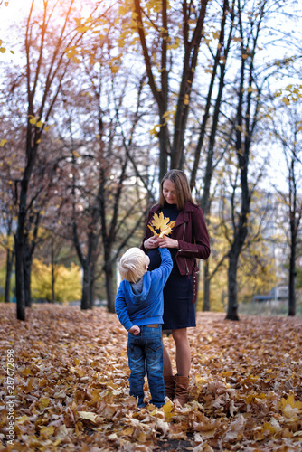 Little blond boy gives his pregnant mother yellow leaf. Autumn park on the background