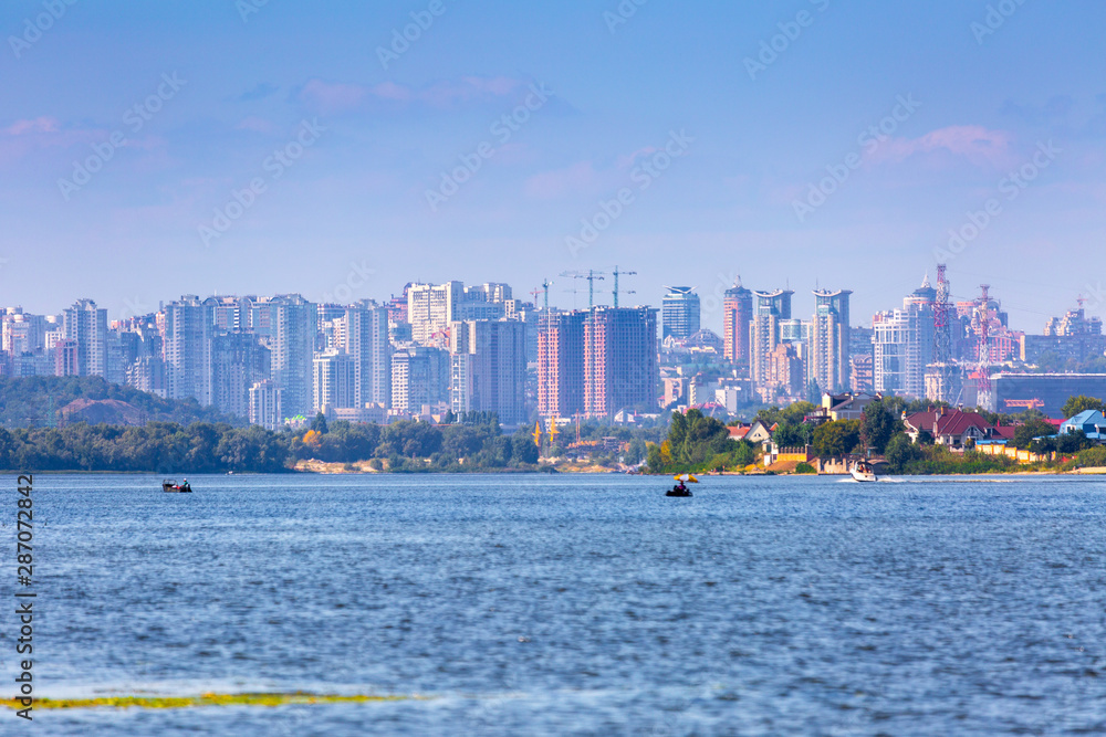 Summer sunny hot evening. Modern residential areas southern suburbs and midtown on hills in Kyiv on the right bank of the Dnipro River. Fishermen on boats on foreground. Kyiv. Ukraine. Aug. 28, 2019