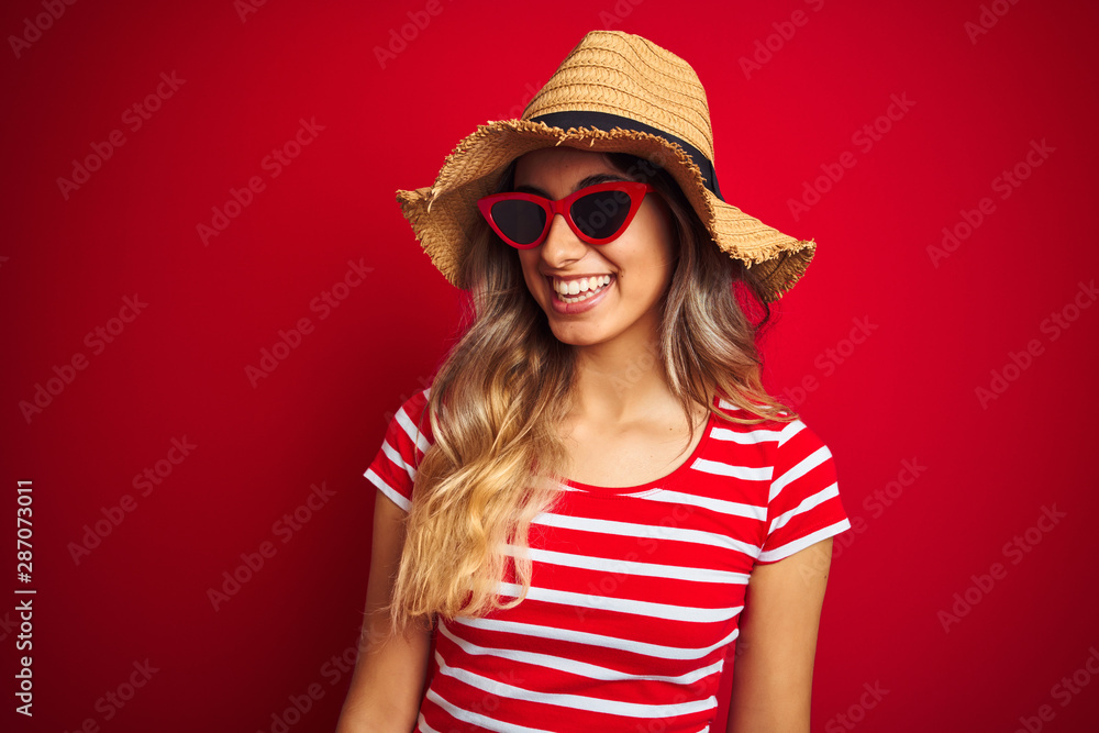Young beautiful woman wearing sunglasses and summer hat over red isolated background looking away to side with smile on face, natural expression. Laughing confident. Young beautiful woman wearing sunglasses and summer hat over red isolated background looking away to side with smile on face, natural expression. Laughing confident.