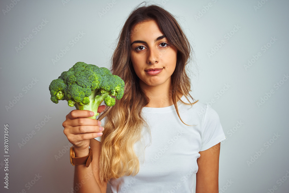 Young beautiful woman eating broccoli over grey isolated background with a confident expression on smart face thinking serious Young beautiful woman eating broccoli over grey isolated background with a confident expression on smart face thinking serious