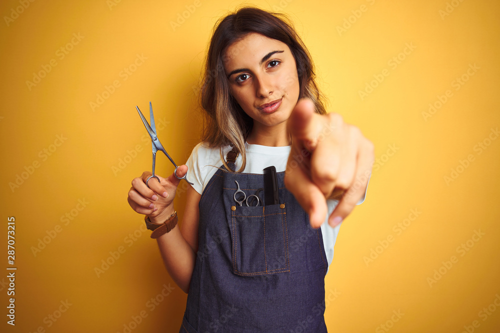 Young beautiful hairdresser woman holding scissors over yellow isolated background pointing with finger to the camera and to you, hand sign, positive and confident gesture from the front Young beautiful hairdresser woman holding scissors over yellow isolated background pointing with finger to the camera and to you, hand sign, positive and confident gesture from the front