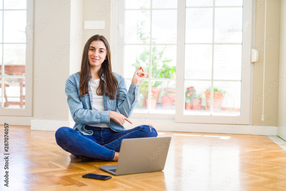 Beautiful young woman sitting on the floor with crossed legs using laptop with a big smile on face, pointing with hand and finger to the side looking at the camera. Beautiful young woman sitting on the floor with crossed legs using laptop with a big smile on face, pointing with hand and finger to the side looking at the camera.