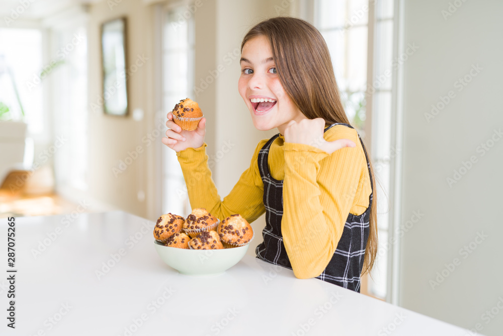 Beautiful young girl eating chocolate chips muffins pointing and showing with thumb up to the side with happy face smiling Beautiful young girl eating chocolate chips muffins pointing and showing with thumb up to the side with happy face smiling