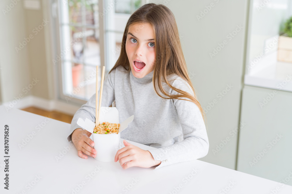 Beautiful young girl kid eating asian rice in delivery box scared in shock with a surprise face, afraid and excited with fear expression Beautiful young girl kid eating asian rice in delivery box scared in shock with a surprise face, afraid and excited with fear expression