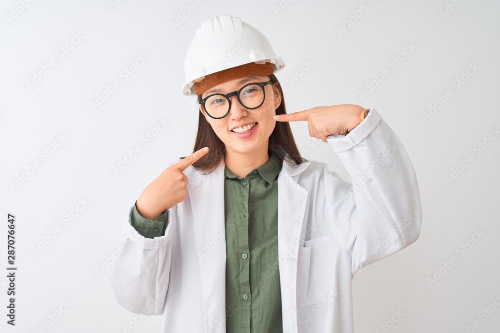 Young chinese engineer woman wearing coat helmet glasses over isolated white background smiling cheerful showing and pointing with fingers teeth and mouth. Dental health concept. Young chinese engineer woman wearing coat helmet glasses over isolated white background smiling cheerful showing and pointing with fingers teeth and mouth. Dental health concept.