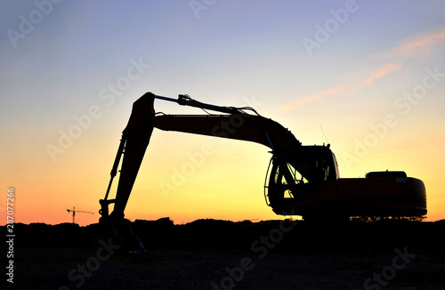 Silhouette of the heavy tracked excavator at a construction site on a background sunset. Special heavy construction equipment for road construction. Small roughness sharpness, possible granularity.