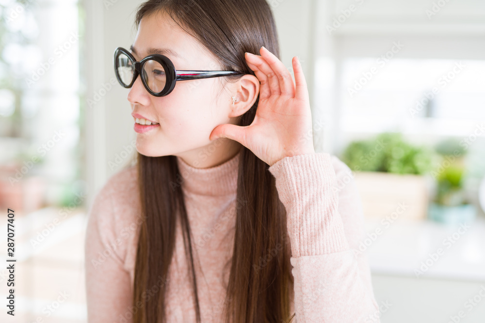 Beautiful Asian woman wearing glasses smiling with hand over ear listening an hearing to rumor or gossip. Deafness concept. Beautiful Asian woman wearing glasses smiling with hand over ear listening an hearing to rumor or gossip. Deafness concept.