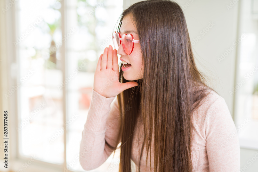 Naklejka premium Beautiful Asian woman wearing pink heart glasses shouting and screaming loud to side with hand on mouth. Communication concept.
