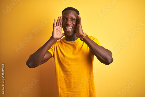 Young african american man wearing casual t-shirt standing over isolated yellow background Smiling cheerful playing peek a boo with hands showing face. Surprised and exited