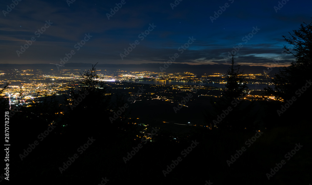 Fototapeta premium Evening top view of city lights, French Annemasse, Swiss Geneva,lake Geneva and picturesque sky with dark clouds after sunset,photo with long exposure.Department of Haute-Savoie in France.