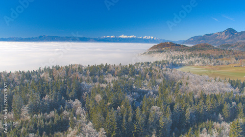 AERIAL: High snowy mountains and church behind icy frozen forest in foggy winter