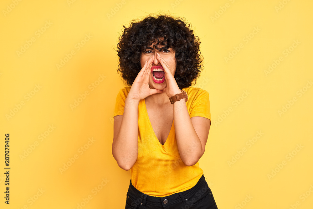 Young arab woman with curly hair wearing t-shirt standing over isolated yellow background Shouting angry out loud with hands over mouth Young arab woman with curly hair wearing t-shirt standing over isolated yellow background Shouting angry out loud with hands over mouth