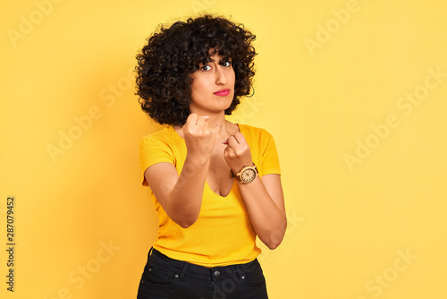 Young arab woman with curly hair wearing t-shirt standing over isolated yellow background Ready to fight with fist defense gesture, angry and upset face, afraid of problem