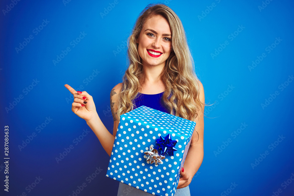 Young beautiful woman holding birthday present over blue isolated background very happy pointing with hand and finger to the side Young beautiful woman holding birthday present over blue isolated background very happy pointing with hand and finger to the side