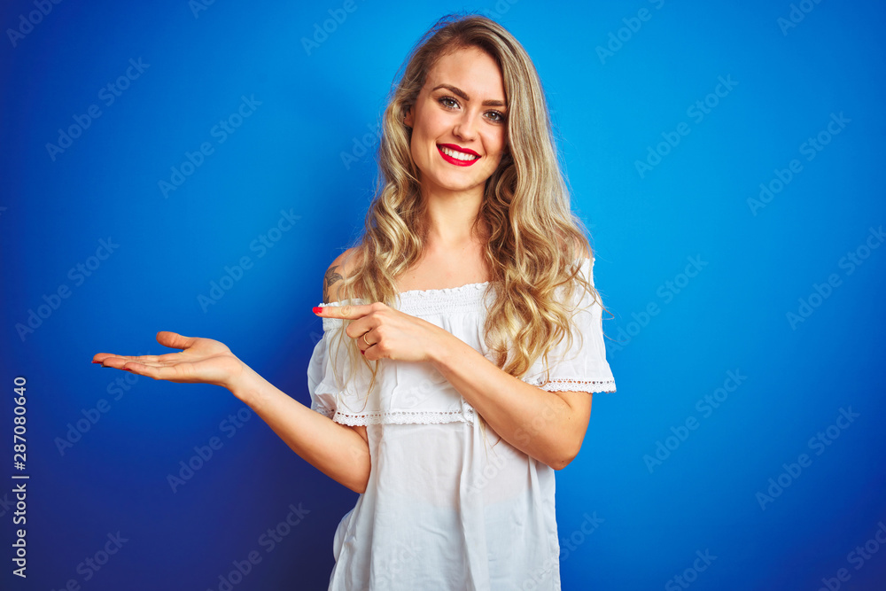 Young beautiful woman wearing white dress standing over blue isolated background amazed and smiling to the camera while presenting with hand and pointing with finger. Young beautiful woman wearing white dress standing over blue isolated background amazed and smiling to the camera while presenting with hand and pointing with finger.