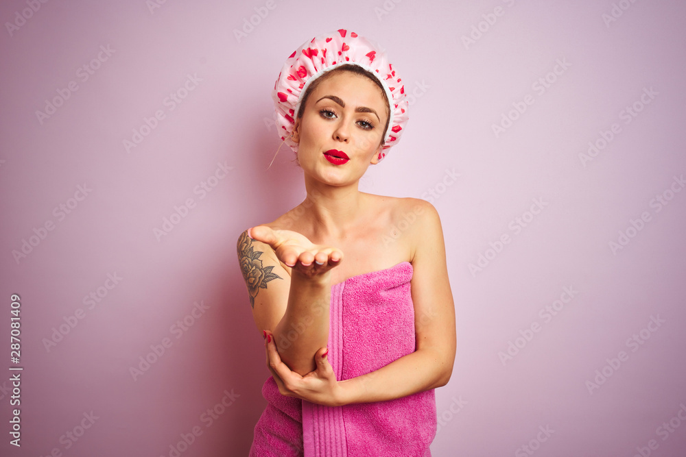 Young beautiful woman wearing towel and bath hat after shower over pink isolated background looking at the camera blowing a kiss with hand on air being lovely and sexy. Love expression. Young beautiful woman wearing towel and bath hat after shower over pink isolated background looking at the camera blowing a kiss with hand on air being lovely and sexy. Love expression.