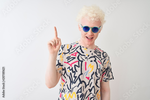 Young albino blond man wearing colorful t-shirt and sunglasses over isolated red background pointing finger up with successful idea. Exited and happy. Number one. © Krakenimages.com
