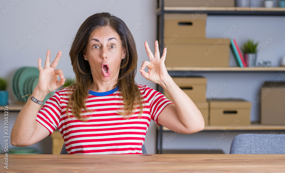 Middle age senior woman sitting at the table at home looking surprised and shocked doing ok approval symbol with fingers. Crazy expression Middle age senior woman sitting at the table at home looking surprised and shocked doing ok approval symbol with fingers. Crazy expression