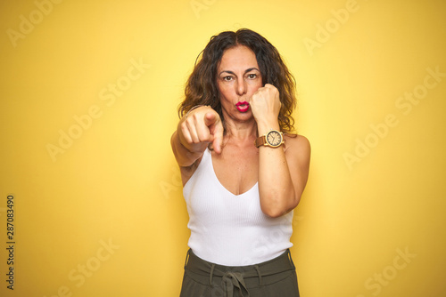 Middle age senior woman showing wrist watch over yellow isolated background pointing with finger to the camera and to you, hand sign, positive and confident gesture from the front