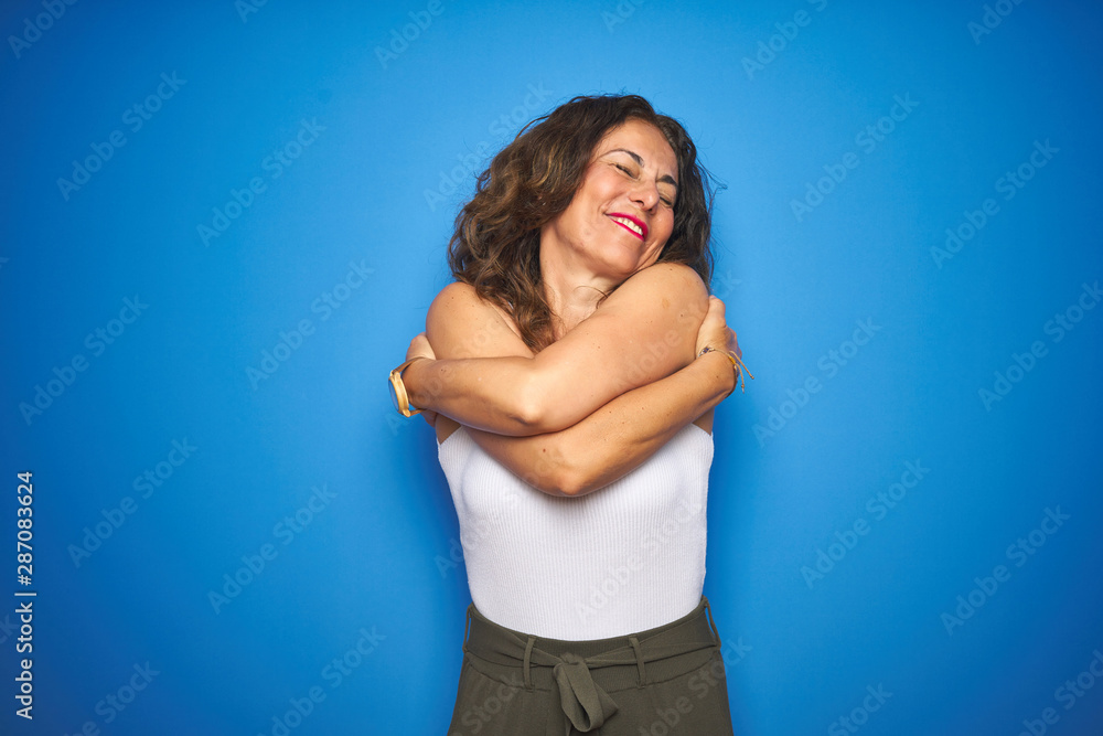 Middle age senior woman with curly hair standing over blue isolated background Hugging oneself happy and positive, smiling confident. Self love and self care Middle age senior woman with curly hair standing over blue isolated background Hugging oneself happy and positive, smiling confident. Self love and self care