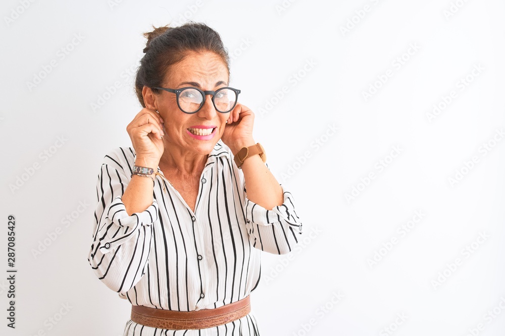 Middle age businesswoman wearing striped dress and glasses over isolated white background covering ears with fingers with annoyed expression for the noise of loud music. Deaf concept. Middle age businesswoman wearing striped dress and glasses over isolated white background covering ears with fingers with annoyed expression for the noise of loud music. Deaf concept.