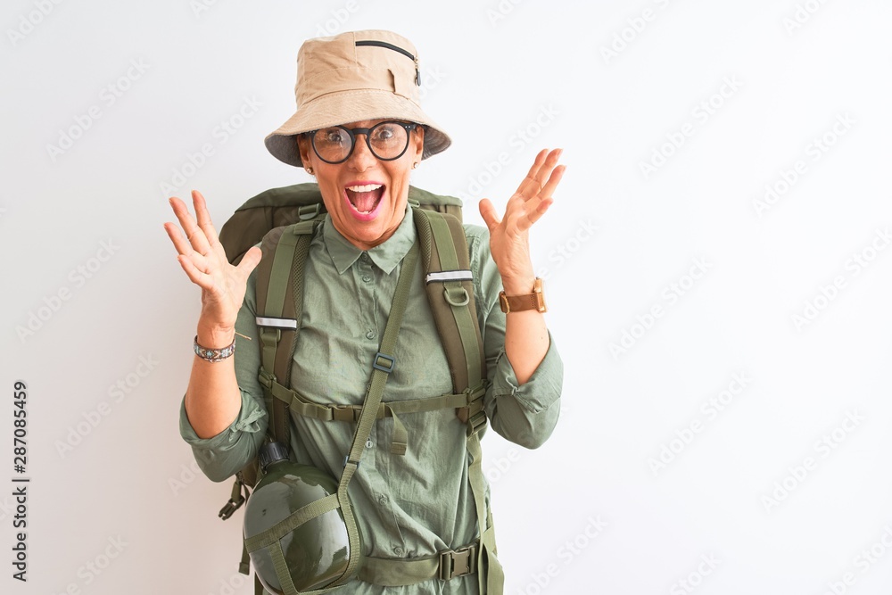 Middle age hiker woman wearing backpack canteen hat glasses over isolated white background celebrating crazy and amazed for success with arms raised and open eyes screaming excited. Winner concept