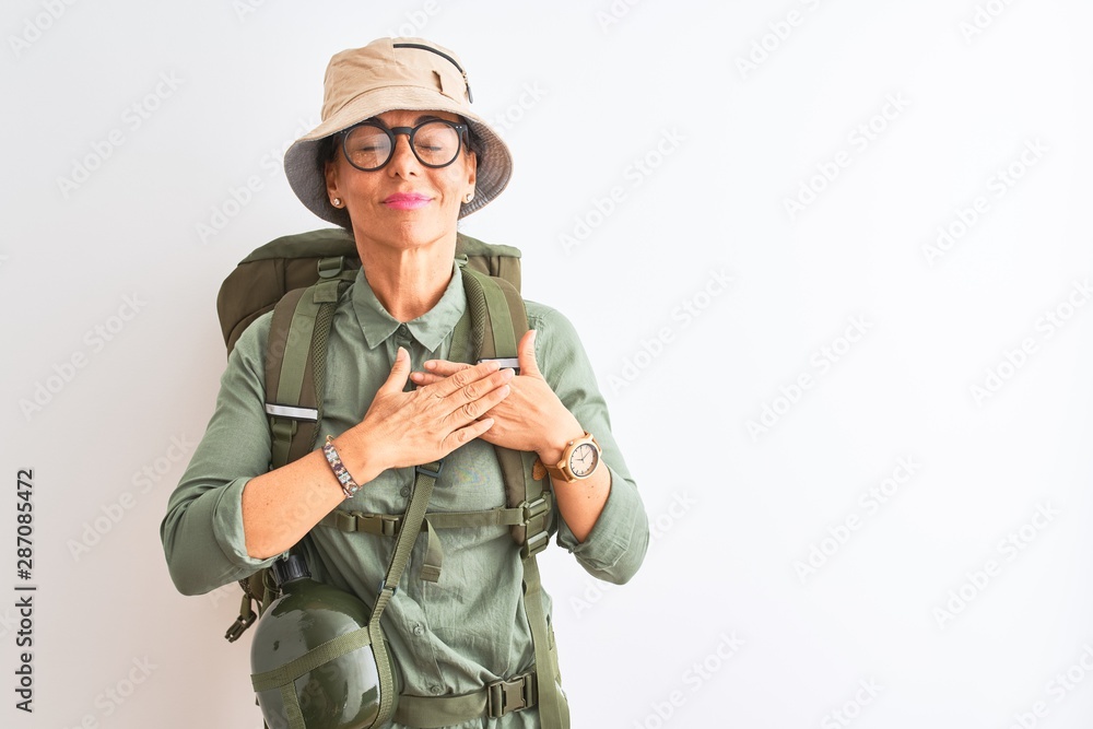 Middle age hiker woman wearing backpack canteen hat glasses over isolated white background smiling with hands on chest with closed eyes and grateful gesture on face. Health concept. Middle age hiker woman wearing backpack canteen hat glasses over isolated white background smiling with hands on chest with closed eyes and grateful gesture on face. Health concept.