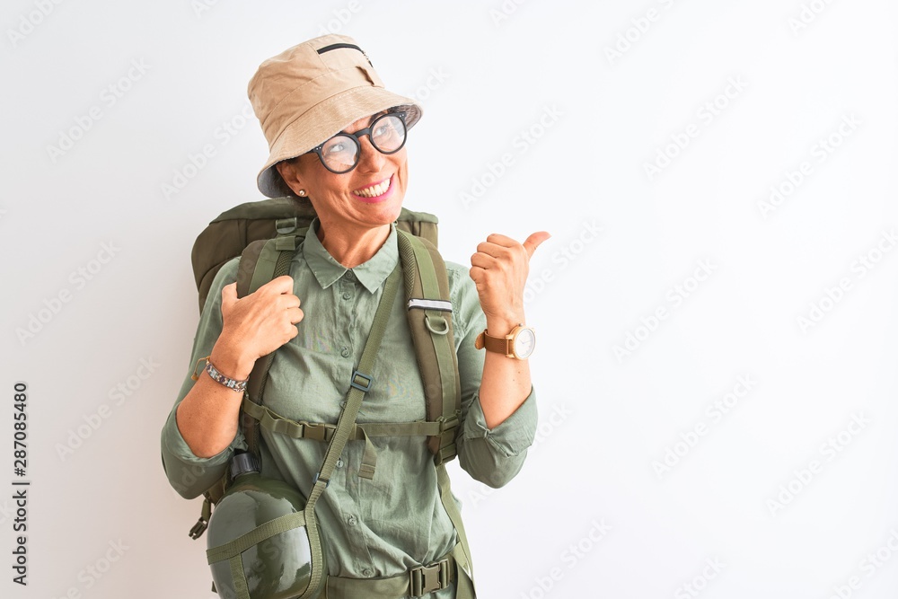 Fototapeta premium Middle age hiker woman wearing backpack canteen hat glasses over isolated white background smiling with happy face looking and pointing to the side with thumb up.