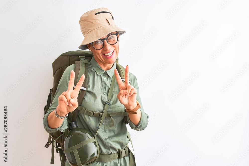 Fototapeta premium Middle age hiker woman wearing backpack canteen hat glasses over isolated white background smiling looking to the camera showing fingers doing victory sign. Number two.