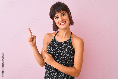 Young beautiful woman wearing black floral dress standing over isolated pink background smiling and looking at the camera pointing with two hands and fingers to the side. © Krakenimages.com