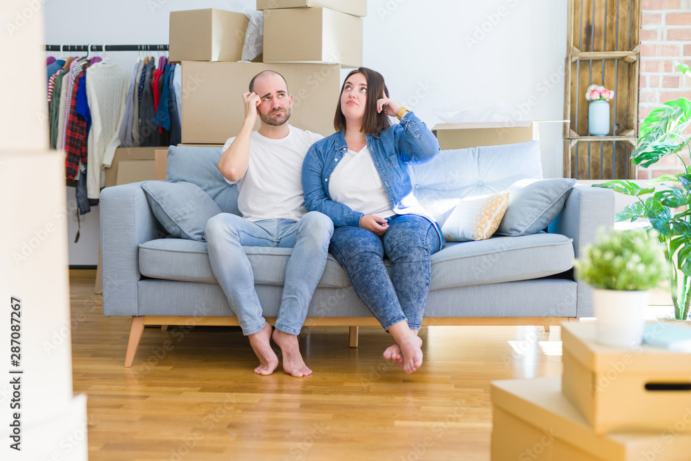 Young couple sitting on the sofa arround cardboard boxes moving to a new house confuse and wonder about question. Uncertain with doubt, thinking with hand on head. Pensive concept. Young couple sitting on the sofa arround cardboard boxes moving to a new house confuse and wonder about question. Uncertain with doubt, thinking with hand on head. Pensive concept.
