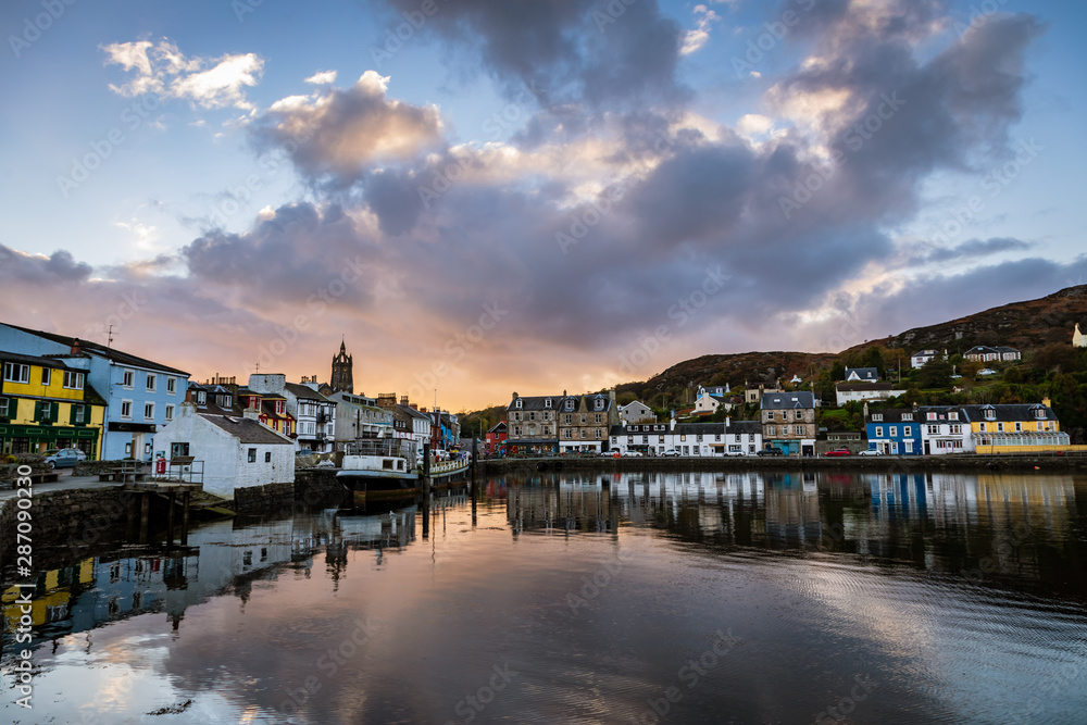 Clouds start to turn pink reflecting in the still water of Tarbert Harbour with the beautifully painted houses on Barmore Rd in Argyll and Bute Scotland United Kingdom