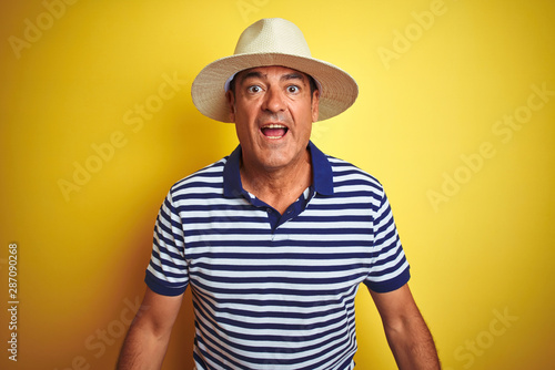 Handsome middle age man wearing striped polo and hat over isolated yellow background afraid and shocked with surprise expression, fear and excited face.