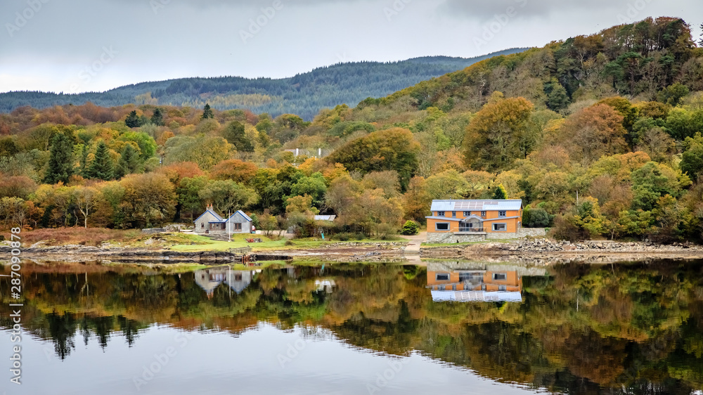 Glassy water reflects two houses in front of fluffy rusty orange and green fall colored trees seen from the Calmac Ferry near  Dunmore and Kennacraig Scotland