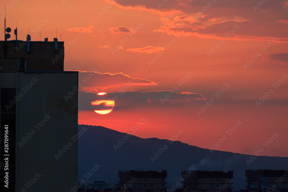 Sunrise in city with red sky, clouds, silhouettes of buildings and mountain. 