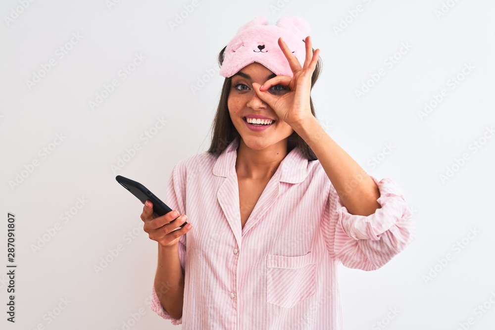 Young woman wearing sleep mask pajama using smartphone over isolated white background with happy face smiling doing ok sign with hand on eye looking through fingers Young woman wearing sleep mask pajama using smartphone over isolated white background with happy face smiling doing ok sign with hand on eye looking through fingers