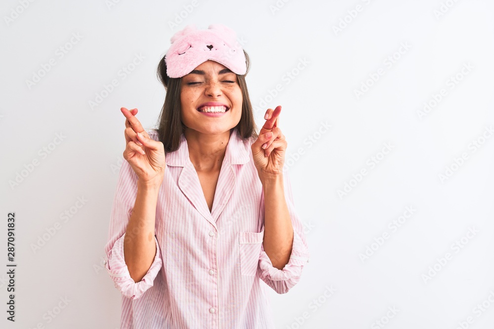 Young beautiful woman wearing sleep mask and pajama over isolated white background gesturing finger crossed smiling with hope and eyes closed. Luck and superstitious concept. Young beautiful woman wearing sleep mask and pajama over isolated white background gesturing finger crossed smiling with hope and eyes closed. Luck and superstitious concept.