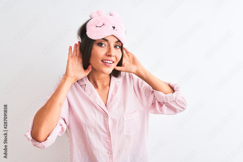 Young beautiful woman wearing sleep mask and pajama over isolated white background Trying to hear both hands on ear gesture, curious for gossip. Hearing problem, deaf Young beautiful woman wearing sleep mask and pajama over isolated white background Trying to hear both hands on ear gesture, curious for gossip. Hearing problem, deaf