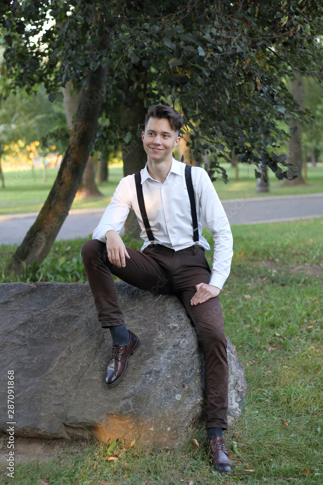 Stylishly dressed young man sits on a stone boulder in the park. With a thoughtful look. Stylishly dressed young man sits on a stone boulder in the park. With a thoughtful look.