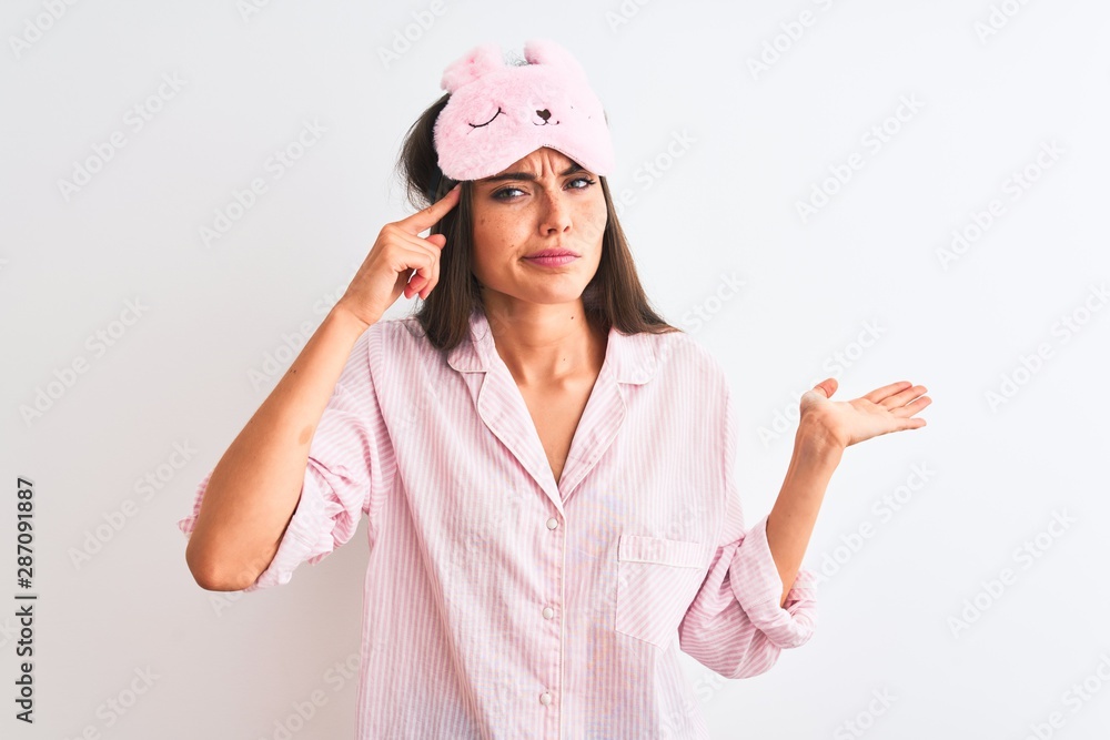 Young beautiful woman wearing sleep mask and pajama over isolated white background confused and annoyed with open palm showing copy space and pointing finger to forehead. Think about it. Young beautiful woman wearing sleep mask and pajama over isolated white background confused and annoyed with open palm showing copy space and pointing finger to forehead. Think about it.