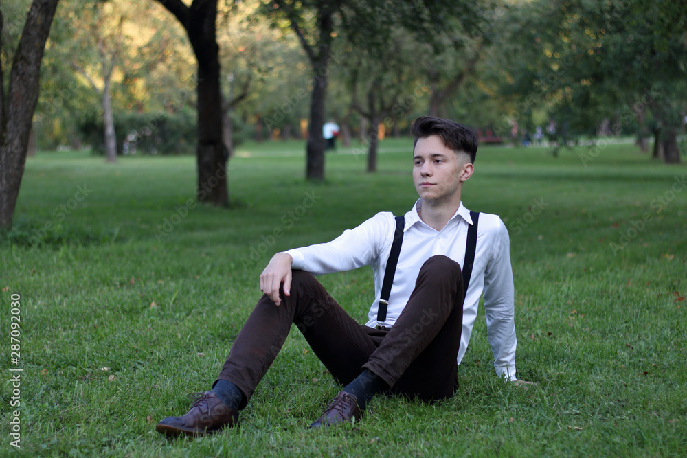 Stylishly dressed young man posing in a park. Sits on the lawn and looks away. Stylishly dressed young man posing in a park. Sits on the lawn and looks away.