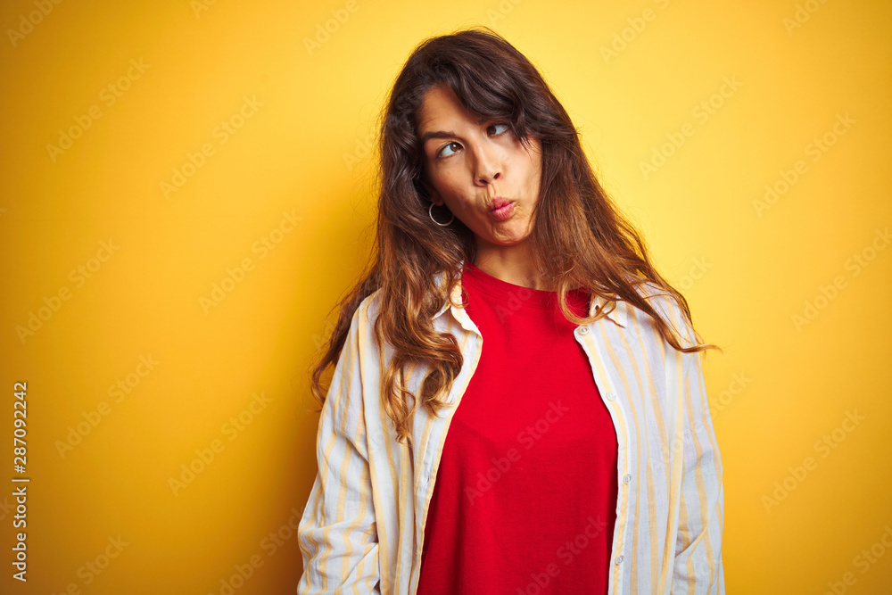 Young beautiful woman wearing red t-shirt and stripes shirt over yellow isolated background making fish face with lips, crazy and comical gesture. Funny expression. Young beautiful woman wearing red t-shirt and stripes shirt over yellow isolated background making fish face with lips, crazy and comical gesture. Funny expression.