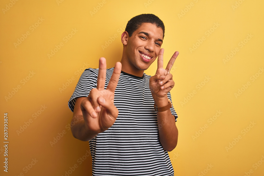 Young handsome arab man wearing navy striped t-shirt over isolated yellow background smiling looking to the camera showing fingers doing victory sign. Number two.