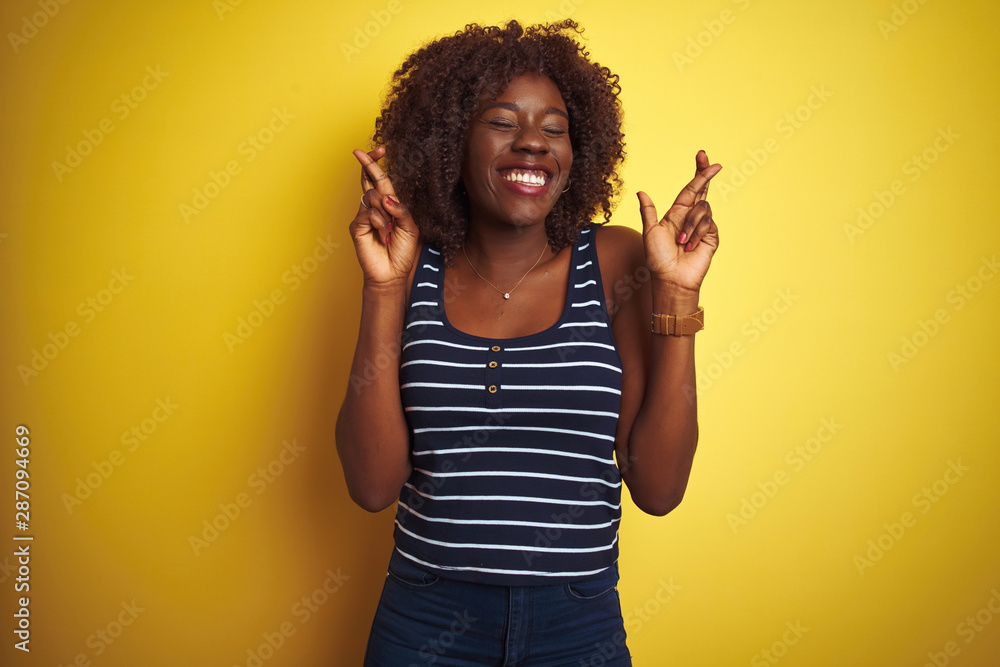 Young african afro woman wearing striped t-shirt over isolated yellow background gesturing finger crossed smiling with hope and eyes closed. Luck and superstitious concept. Young african afro woman wearing striped t-shirt over isolated yellow background gesturing finger crossed smiling with hope and eyes closed. Luck and superstitious concept.