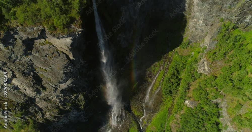 Aerial: Rainbow appearing over misty waterfall falling from rocky cliff on moss covered land during sunny day - Geiranger Fjord, Norway Aerial: Rainbow appearing over misty waterfall falling from rocky cliff on moss covered land during sunny day - Geiranger Fjord, Norway