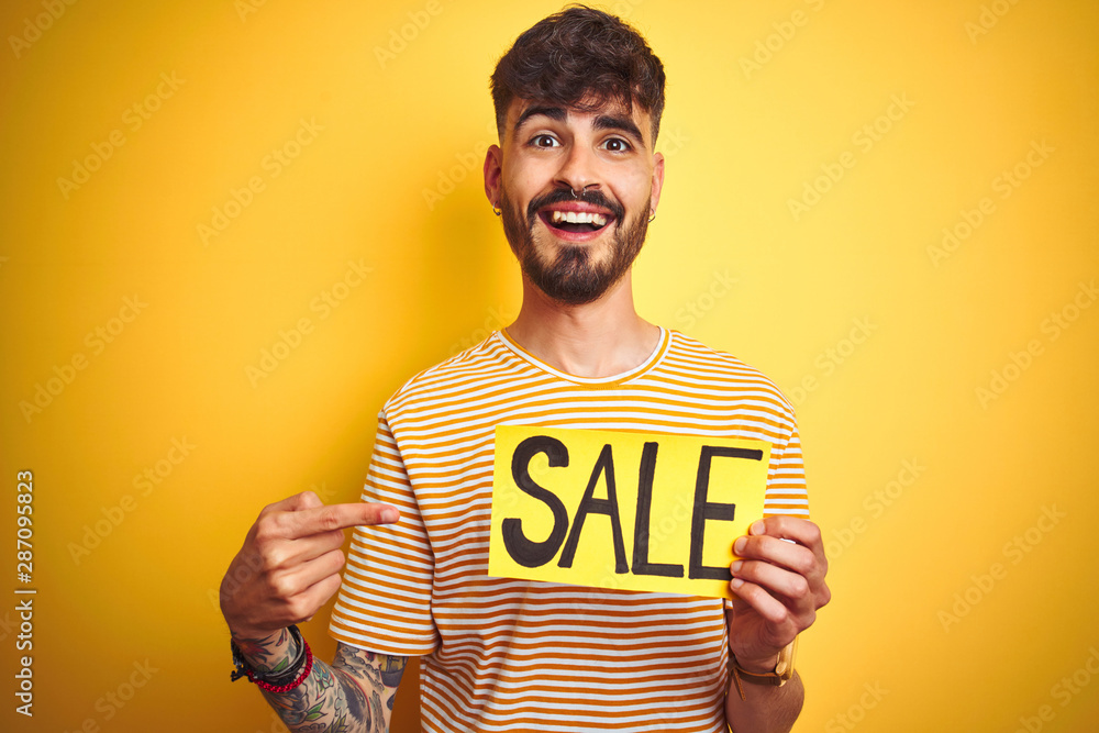 Young man with tattoo holding sale banner standing over isolated yellow background very happy pointing with hand and finger Young man with tattoo holding sale banner standing over isolated yellow background very happy pointing with hand and finger