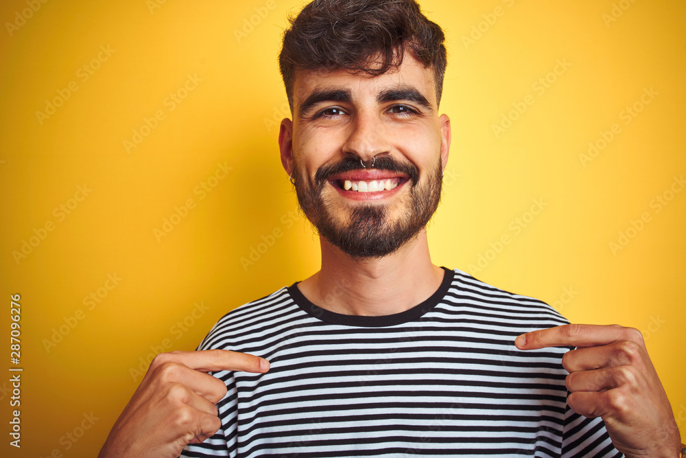 Young man with tattoo wearing striped t-shirt standing over isolated yellow background looking confident with smile on face, pointing oneself with fingers proud and happy. Young man with tattoo wearing striped t-shirt standing over isolated yellow background looking confident with smile on face, pointing oneself with fingers proud and happy.