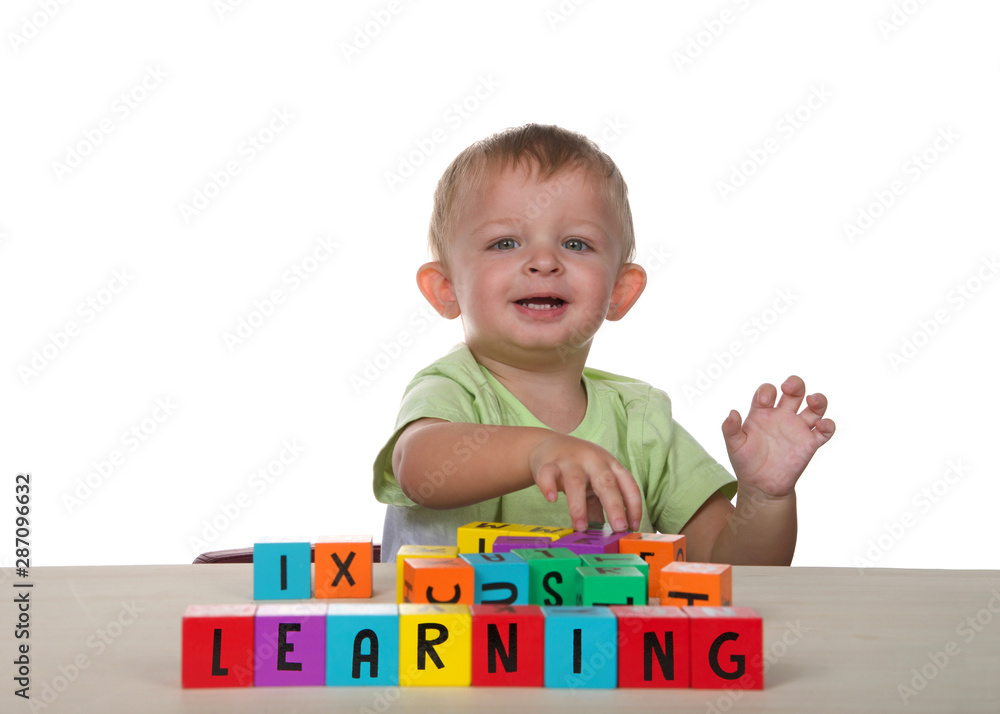 Caucasian toddler male child playing learning with colorful wooden lettered blocks on light wood table, looking towards viewer with happy expression. Isolated on white background. Caucasian toddler male child playing learning with colorful wooden lettered blocks on light wood table, looking towards viewer with happy expression. Isolated on white background.