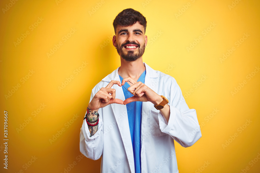 Young doctor man with tattoo standing over isolated yellow background smiling in love showing heart symbol and shape with hands. Romantic concept. Young doctor man with tattoo standing over isolated yellow background smiling in love showing heart symbol and shape with hands. Romantic concept.