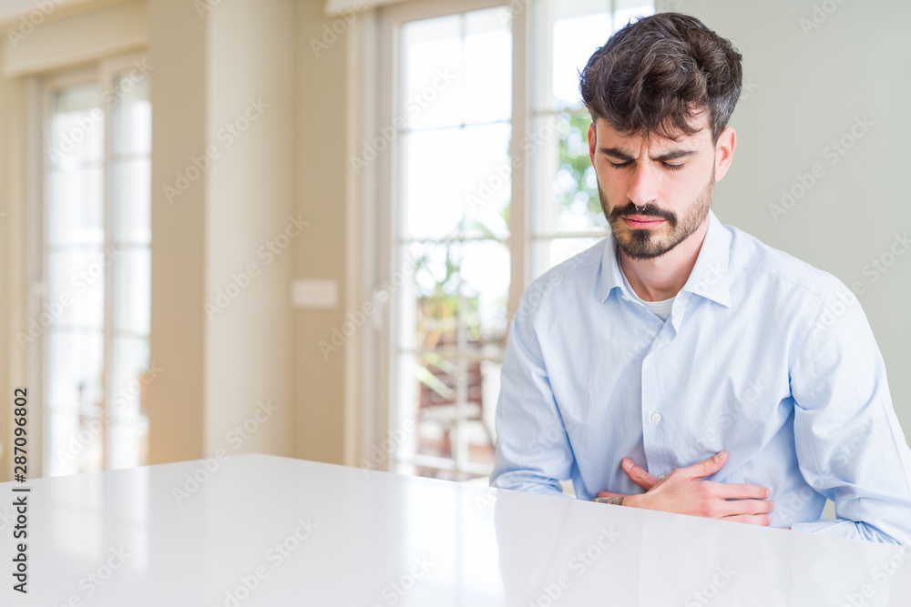 Young businesss man sitting on white table with hand on stomach because nausea, painful disease feeling unwell. Ache concept.